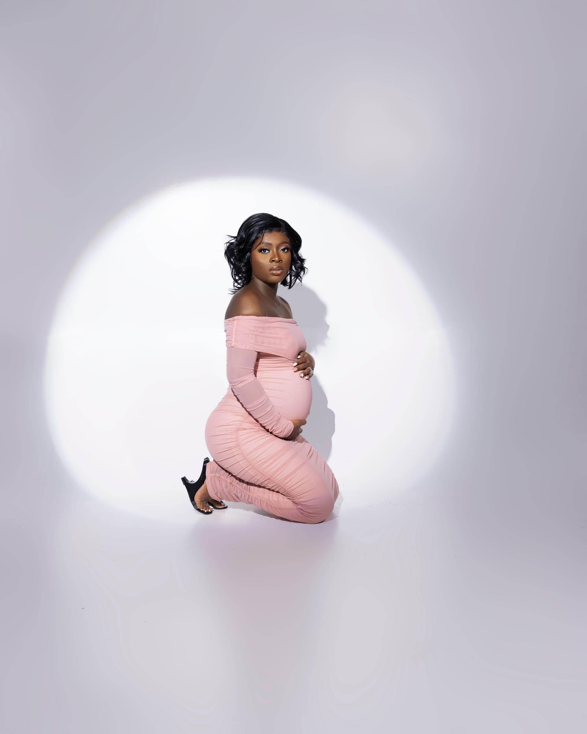 Studio maternity portrait with a subject kneeling in a soft spotlight.